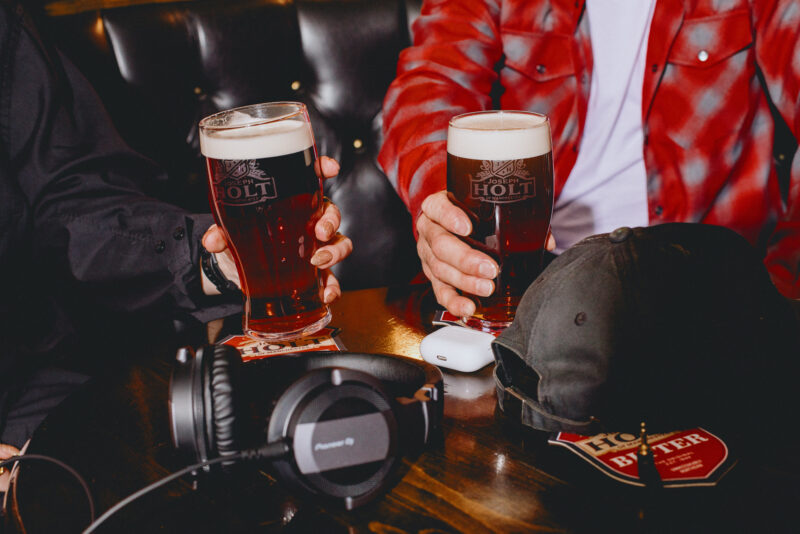 Two pints of bitter next to some headphones, airpods and a hat